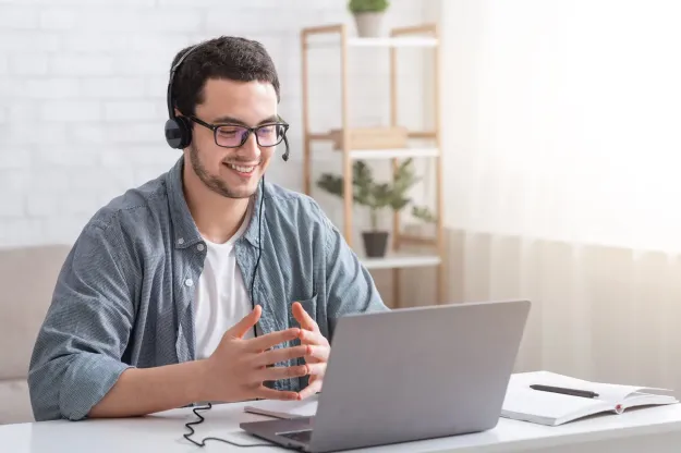Man wearing a headset and glasses, smiling while using a laptop at a desk in an office setting.